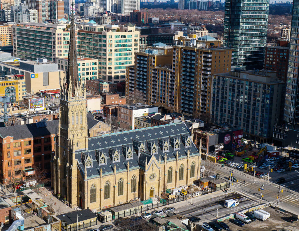 A brick church with a tall steeple surrounded by tall skyscrapers in a busy downtown area