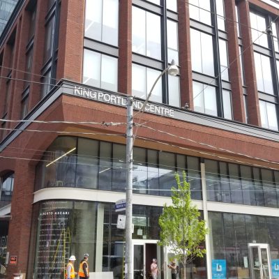 Front of a red brick building on a busy street with large windows and a curved storefront window