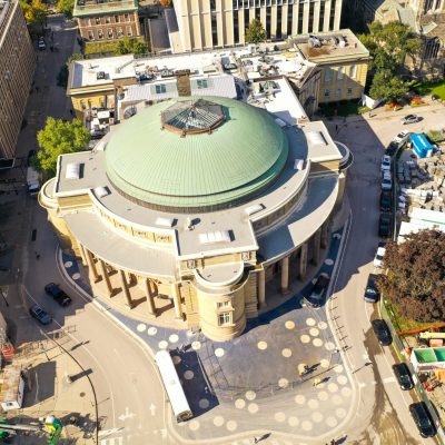 Aerial view of a round building with a green copper roof, construction fencing and mahinery surrounding ther building