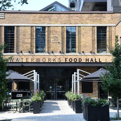 Exterior of beige building with tall windows on the second story with a sign that says waterworks food hall. With greenery and a patio out front