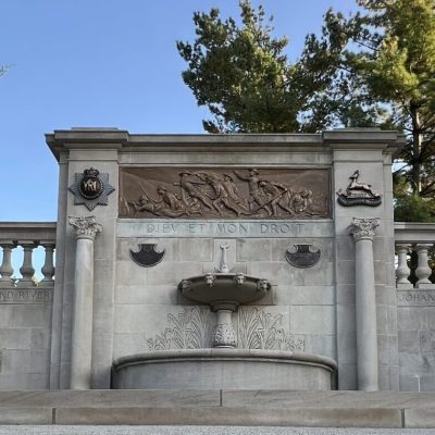 A fountain made of sone with stone walls on either side and a blue sky with trees in the background